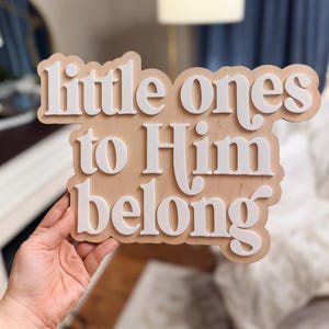 May include: Wooden sign with the phrase "little ones to Him belong" in white, layered lettering. The sign has a light brown background and is held by a person. The sign is likely for a nursery or children's room.