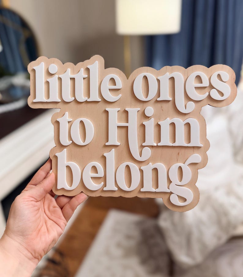 May include: Wooden sign with the phrase "little ones to Him belong" in white, layered lettering. The sign has a light brown background and is held by a person. The sign is likely for a nursery or children's room.