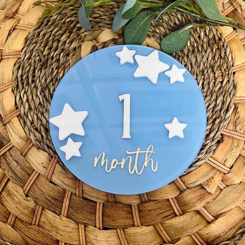 May include: A light blue, round milestone marker with white stars and the number "1". The word "month" is written in gold script below the number. The marker is placed on a woven, natural-colored surface.