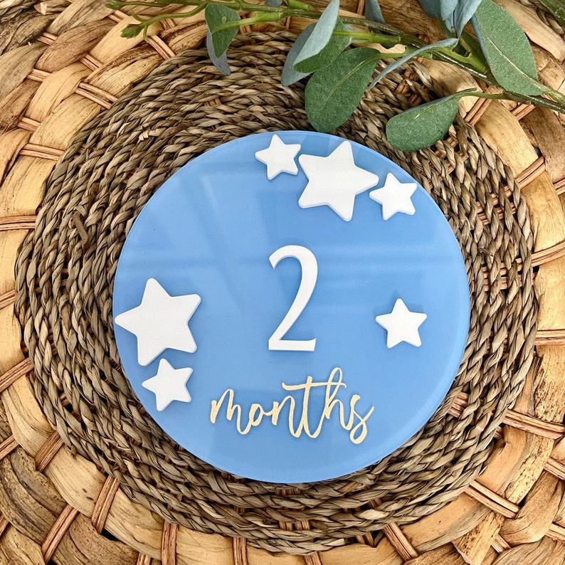 May include: A light blue, round baby milestone plaque with white star accents. The number "2" and the word "months" are displayed in white and gold lettering. The plaque is set on a woven, circular placemat with greenery.