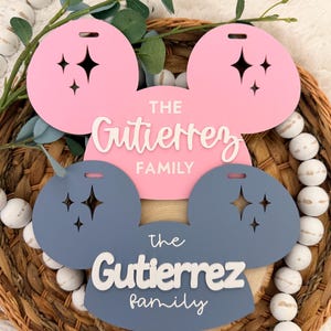 May include: A pink and blue family name sign in the shape of a mouse head. The sign has the name "Gutierrez Family" in white script. The ears have star cutouts. The sign is displayed on a woven basket with white beads.