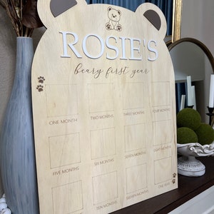 Puede incluir: Un marco de fotos de madera con forma de oso con el nombre "ROSIE'S beary first year" grabado en &eacute;l. El marco tiene 12 espacios para fotos, cada uno etiquetado con un mes del a&ntilde;o.