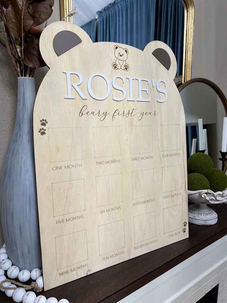 Puede incluir: Un marco de fotos de madera con forma de oso con el nombre "ROSIE'S beary first year" grabado en &eacute;l. El marco tiene 12 espacios para fotos, cada uno etiquetado con un mes del a&ntilde;o.