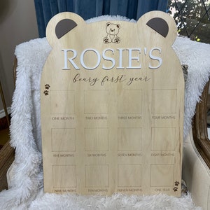 Puede incluir: Un gr&aacute;fico de crecimiento de madera en forma de oso con el nombre "ROSIE'S beary first year" grabado en &eacute;l. El gr&aacute;fico tiene 12 espacios para fotos, cada uno etiquetado con un mes del a&ntilde;o.