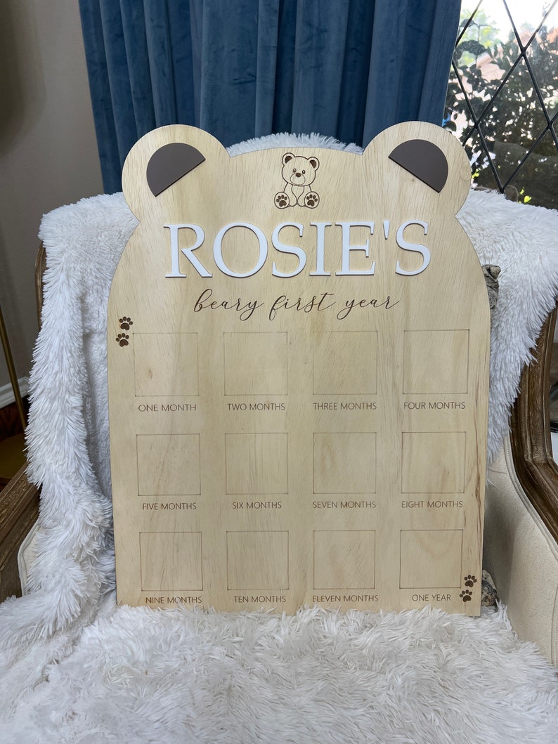 Puede incluir: Un gr&aacute;fico de crecimiento de madera en forma de oso con el nombre "ROSIE'S beary first year" grabado en &eacute;l. El gr&aacute;fico tiene 12 espacios para fotos, cada uno etiquetado con un mes del a&ntilde;o.