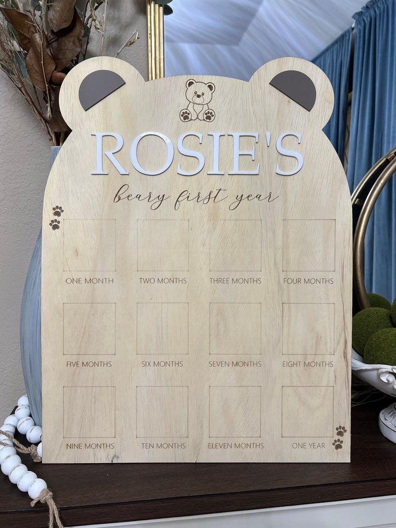 Puede incluir: Un gr&aacute;fico de crecimiento de madera en forma de oso con el nombre "ROSIE'S beary first year" impreso en &eacute;l. El gr&aacute;fico tiene 12 espacios para fotos, cada uno etiquetado con un mes del a&ntilde;o.