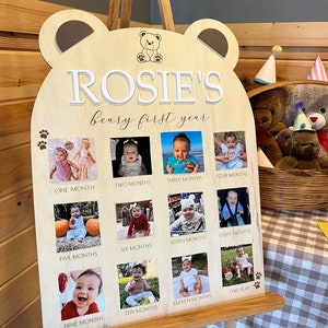 Puede incluir: Un letrero de madera con forma de oso con el nombre "ROSIE'S beary first year" escrito en &eacute;l. El letrero tiene 12 ranuras para fotos, cada una con una foto de un beb&eacute; a diferentes edades, de un mes a un a&ntilde;o.
