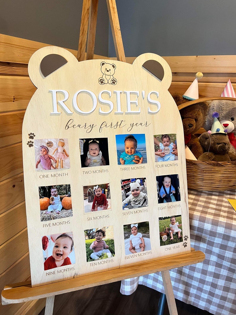 Puede incluir: Un letrero de madera con forma de oso con el nombre "ROSIE'S beary first year" escrito en &eacute;l. El letrero tiene 12 ranuras para fotos, cada una con una foto de un beb&eacute; a diferentes edades, de un mes a un a&ntilde;o.