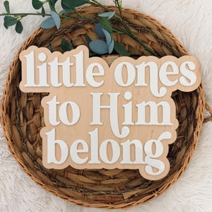 May include: A wooden sign with the words "little ones to Him belong" in white, layered over a light brown wooden base. The sign is placed on a woven basket with greenery.