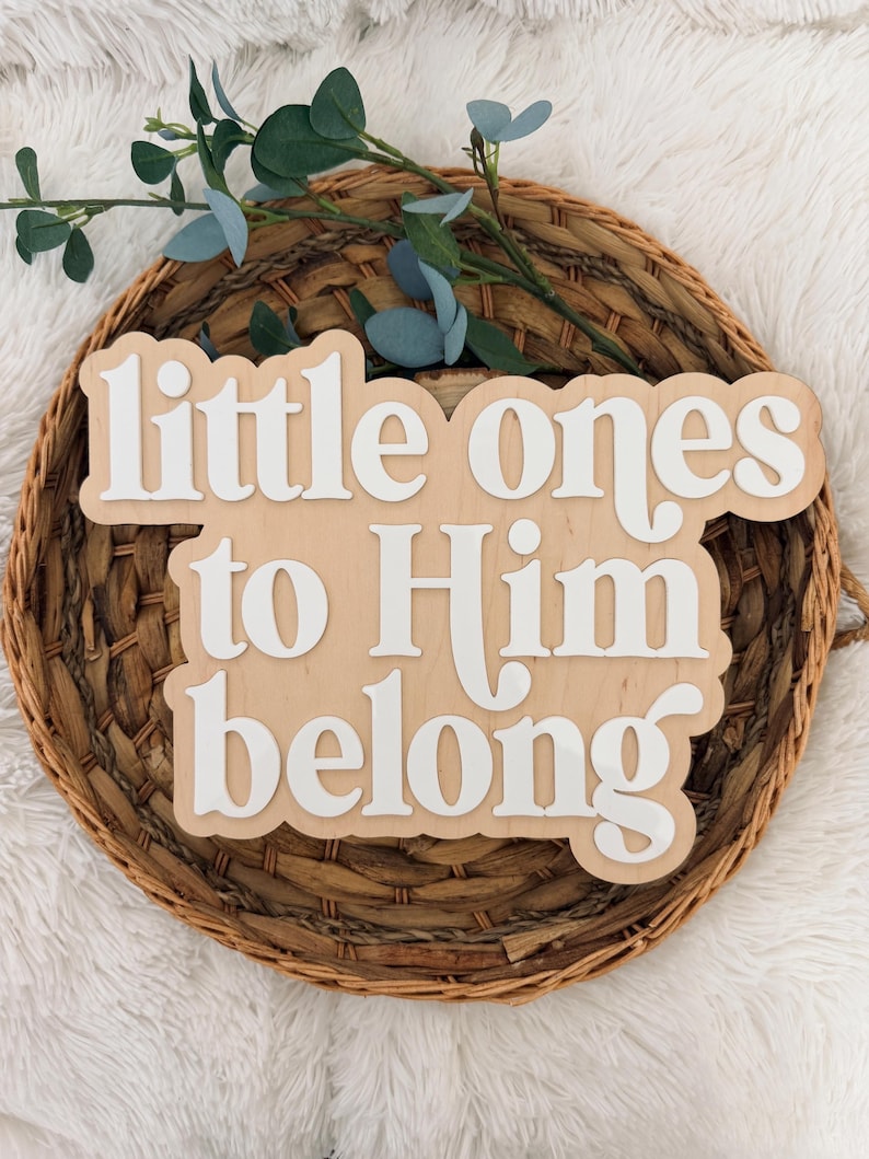 May include: A wooden sign with the words "little ones to Him belong" in white, layered over a light brown wooden base. The sign is placed on a woven basket with greenery.