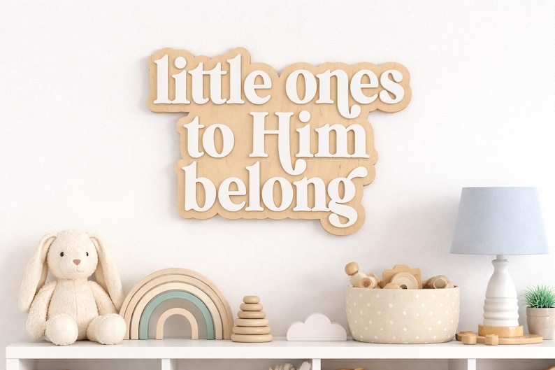 May include: A wooden sign with the words "little ones to Him belong" in white lettering. The sign is above a shelf with a stuffed bunny, a rainbow, a wooden toy, a polka-dot basket, a lamp, and a small plant.