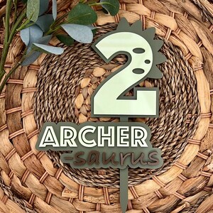 Puede incluir: Topper de pastel de dinosaurio verde y blanco con el n&uacute;mero 2 y el texto "ARCHER -saur-us".