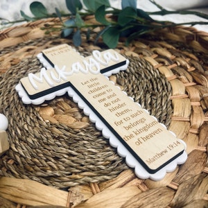 May include: A wooden cross with a white border and scalloped edges. The cross is engraved with the name "Mikayla" and a bible verse: "Let the little children come to me and do not hinder them, for to such belongs the kingdom of heaven Matthew 19:14".