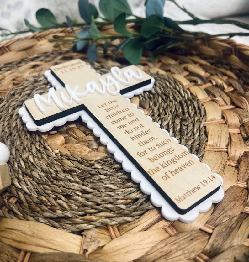 May include: A wooden cross with a white border and scalloped edges. The cross is engraved with the name "Mikayla" and a bible verse: "Let the little children come to me and do not hinder them, for to such belongs the kingdom of heaven Matthew 19:14".