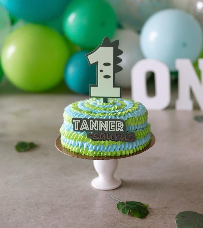 Puede incluir: Un pastel de dos pisos decorado con glaseado verde y azul. El pastel est&aacute; cubierto con un adorno de pastel de dinosaurio en forma de n&uacute;mero uno y un letrero que dice "Tanner-saurus".