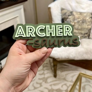 Puede incluir: Pegatina de vinilo 3D verde y blanca con el texto "ARCHER-saurus" en una fuente divertida.