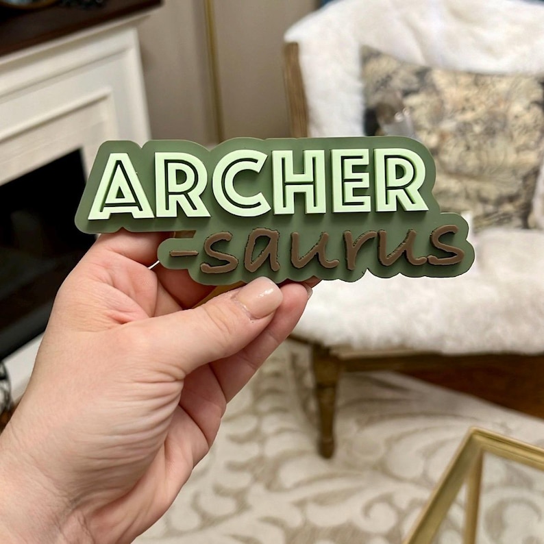Puede incluir: Pegatina de vinilo 3D verde y blanca con el texto "ARCHER-saurus" en una fuente divertida.