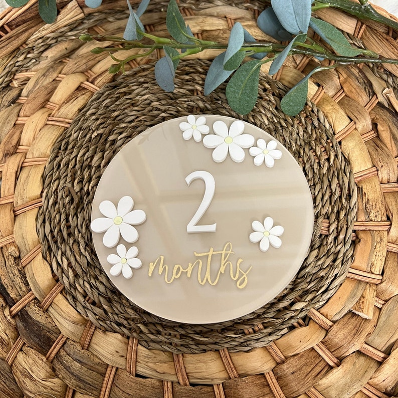 May include: A beige round acrylic milestone marker with white daisy flowers and the number 2 in white with gold writing that says "2 months".