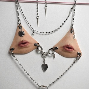 Peut inclure: Un bijou de corps unique en son genre, avec un design de type soutien-gorge orné de triangles couleur peau, chacun orné d'une paire de lèvres. Le bijou est orné de chaînes argentées, de breloques et de pointes, créant une esthétique audacieuse et avant-gardiste.