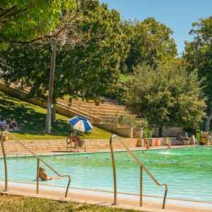 Könnte beinhalten: Ein öffentliches Schwimmbad mit klarem Wasser, umgeben von einer Rasenfläche und Bäumen. Menschen sonnen sich und schwimmen. Eine Rettungsschwimmerstation mit blau-weißem Sonnenschirm ist sichtbar.