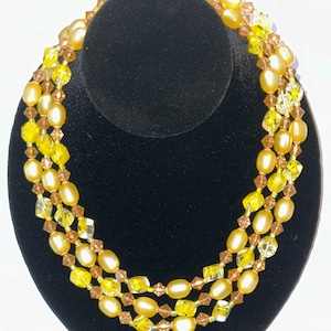 May include: A three-strand necklace with yellow and brown beads. The necklace is made of glass beads and is displayed on a black velvet stand.