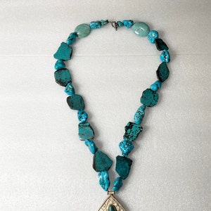 May include: A turquoise gemstone necklace with a silver pendant featuring a turquoise stone. The necklace is made of irregular shaped turquoise stones.
