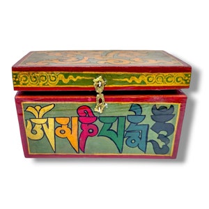 Puede incluir: Una pequeña caja de madera con un diseño pintado en rojo, verde y dorado. La caja tiene una tapa con bisagras y un cierre de latón y está decorada con escritura tibetana colorida.