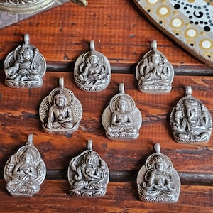 Op de afbeelding: Een set van tien zilveren hangers met verschillende Boeddhistische goden, elk met ingewikkelde details en een traditioneel ontwerp. De hangers worden getoond op een houten oppervlak.