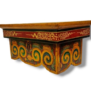Puede incluir: Un estante de madera con intrincados detalles pintados a mano. El estante está pintado en tonos de amarillo, rojo, verde y negro. El estante tiene un borde decorativo con un patrón repetitivo de espirales y flores.