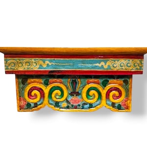 Pode incluir: Uma prateleira de parede retangular de madeira com uma superfície superior plana e um painel frontal decorativo. A prateleira é pintada com cores vibrantes vermelho, amarelo, turquesa e verde, exibindo intrincados motivos florais e volutas.