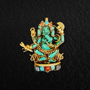 Può includere: Un ciondolo Ganesha verde turchese con dettagli dorati e pietre preziose rosse e blu. Il ciondolo è finemente lavorato e presenta un Ganesha seduto con la proboscide sollevata verso la bocca.