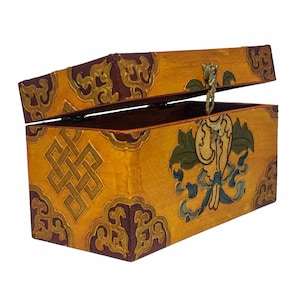 Può includere: Una scatola di legno con un coperchio incernierato, decorata con intricati motivi dorati e rossi. La scatola presenta un design tradizionale di nodo tibetano su un lato e un uccello stilizzato con elementi floreali sull'altro. La scatola ha una chiusura in ottone.