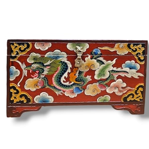 Può includere: Una scatola di legno rossa con un disegno di drago dipinto sul davanti. Il drago è verde, blu e giallo, e la scatola ha accenti dorati. La scatola ha un coperchio incernierato e una chiusura in metallo.