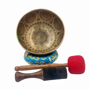 Può includere: Una ciotola tibetana dorata con intricati disegni incisi, appoggiata su un cuscino blu con dettagli dorati. Include un martello di legno con testa in feltro rosso e un piccolo battente di legno con estremità in feltro nero. Ideale per la meditazione e la guarigione sonora.