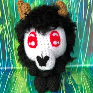 May include: A crocheted plush toy with a black and white skull face, red eyes, and brown horns. The toy is sitting on a green and blue background.