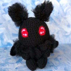 May include: Black crocheted plush toy with large red eyes. The toy is shaped like a moth with fuzzy antennae and wings.