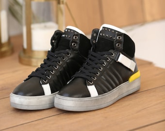 May include: Black high-top trainers with white detailing and a grey sole. The trainers have black laces, a padded collar, and a strap with a silver buckle. The heel has a yellow accent.