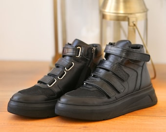 May include: A pair of black high-top trainers with multiple hook-and-loop straps and a zip on one side. The trainers have a mesh toe and a thick, black sole. The shoes are made of leather and are displayed on a wooden surface.