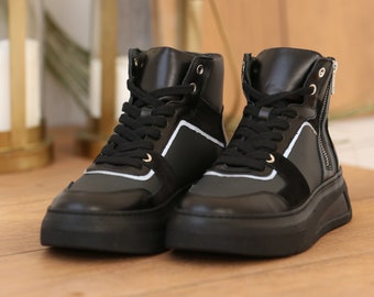 May include: Black high-top trainers with white detailing and a side zip. The trainers feature black laces and a chunky black sole. The shoes are made from leather and suede.