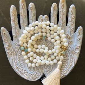 May include: A white mother of pearl mala necklace with green gemstone accents and a gold tassel. The necklace is displayed on a decorative hand-shaped dish.