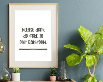 May include: A framed print with a white background and black text that reads "Please don't do coke in our bathroom." The print is hanging on a blue wall with a green plant in a pot to the right.