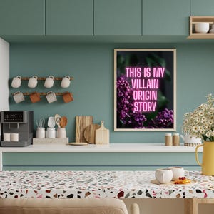 May include: A kitchen scene with a framed print that reads "THIS IS MY VILLAIN ORIGIN STORY" in pink neon. The print is above a countertop with a coffee maker, mugs, and cutting boards. A vase of white flowers and a yellow pitcher are also visible.