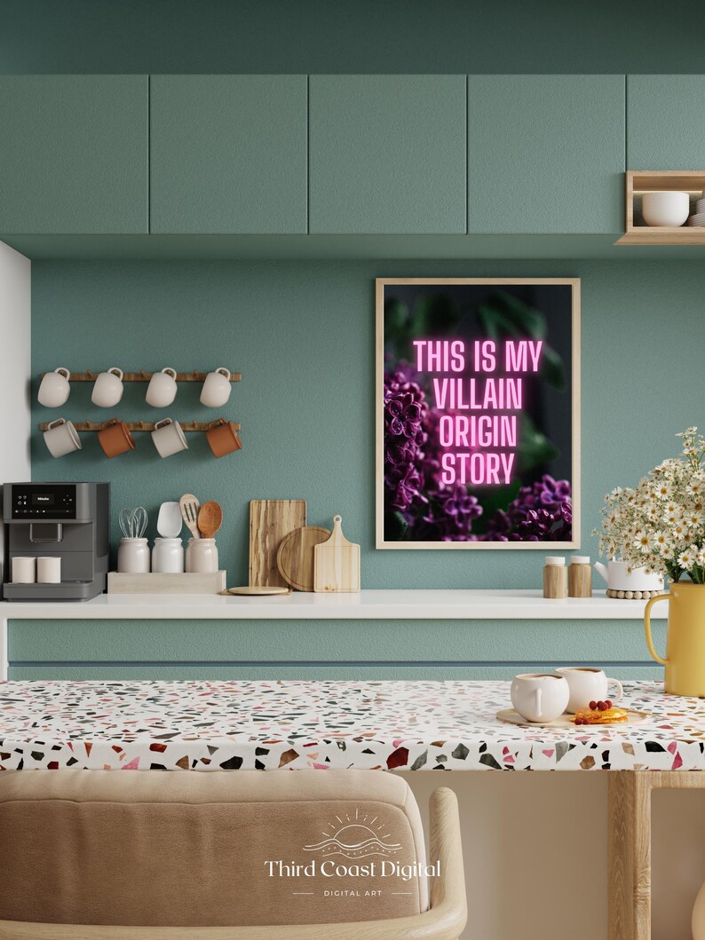 May include: A kitchen scene with a framed print that reads "THIS IS MY VILLAIN ORIGIN STORY" in pink neon. The print is above a countertop with a coffee maker, mugs, and cutting boards. A vase of white flowers and a yellow pitcher are also visible.