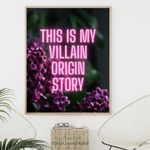 May include: A framed art print with the text "THIS IS MY VILLAIN ORIGIN STORY" in pink neon against a backdrop of purple flowers. The artwork is displayed on a white wall, with a wooden side table and a modern chair in the foreground.