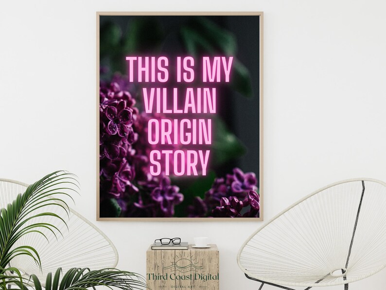 May include: A framed art print with the text "THIS IS MY VILLAIN ORIGIN STORY" in pink neon against a backdrop of purple flowers. The artwork is displayed on a white wall, with a wooden side table and a modern chair in the foreground.