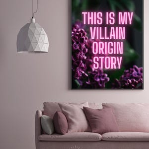 May include: A framed art print with the text "THIS IS MY VILLAIN ORIGIN STORY" in pink neon against a dark background with purple flowers. The print hangs above a pink sofa with pillows.