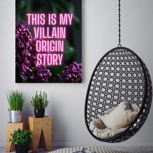 May include: A framed canvas print with the text "THIS IS MY VILLAIN ORIGIN STORY" in pink neon lettering, set against a backdrop of purple flowers. A black hanging chair and small potted plants are also visible.