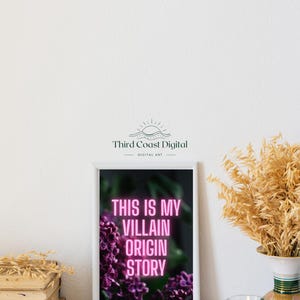 May include: A framed art print with the text "THIS IS MY VILLAIN ORIGIN STORY" in pink neon against a dark background with purple flowers. The print is in a white frame, and the image is set on a white surface with a candle and vase.