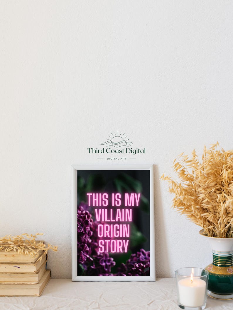 May include: A framed art print with the text "THIS IS MY VILLAIN ORIGIN STORY" in pink neon against a dark background with purple flowers. The print is in a white frame, and the image is set on a white surface with a candle and vase.