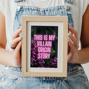 May include: A framed print with the text "THIS IS MY VILLAIN ORIGIN STORY" in pink neon against a dark background with purple flowers. The frame is light brown, and the image is held by a person wearing denim overalls.
